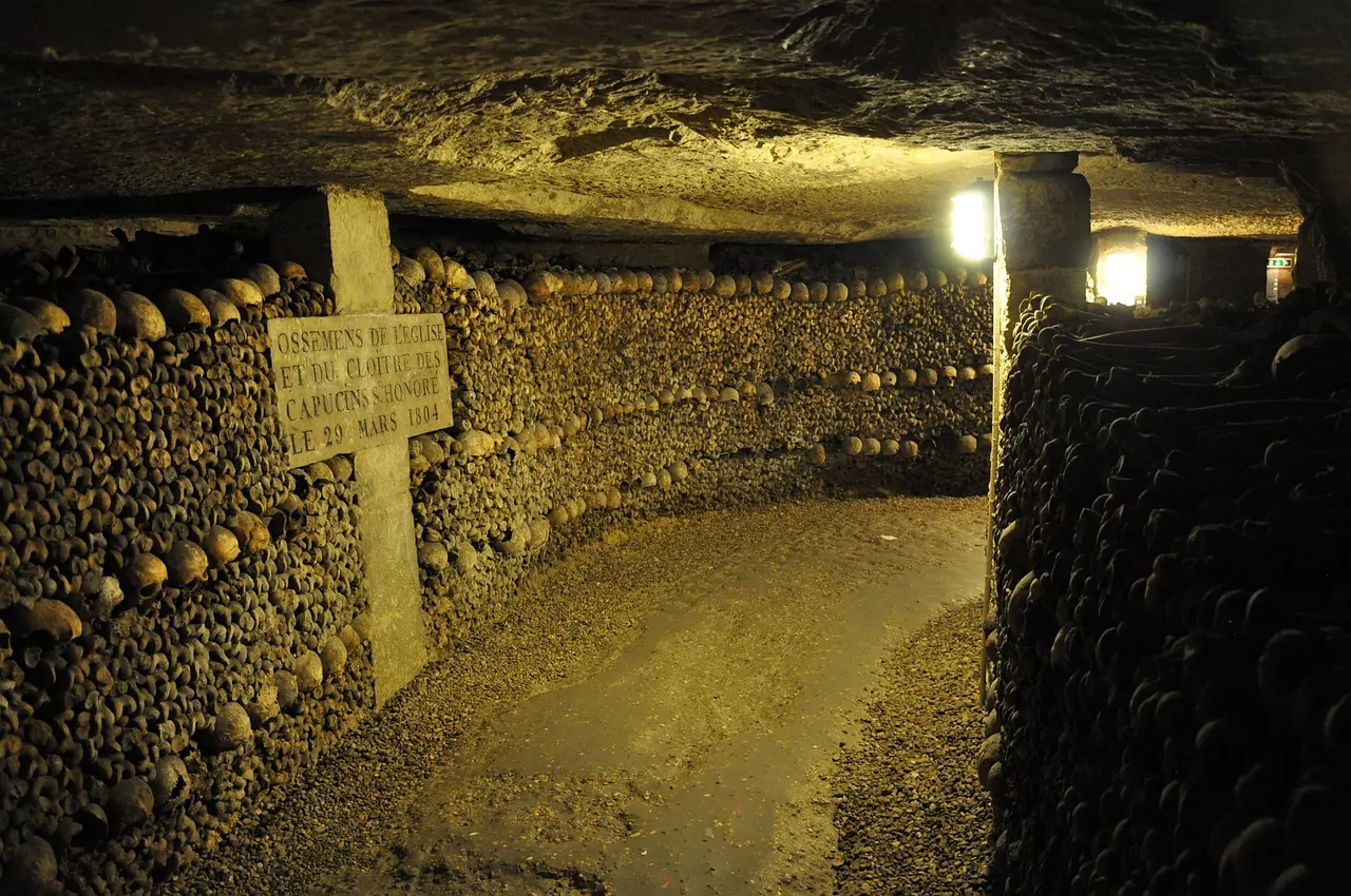 Underground tunnel with walls of human skulls and bones, illuminated by dim light. A stone sign is visible on the left.