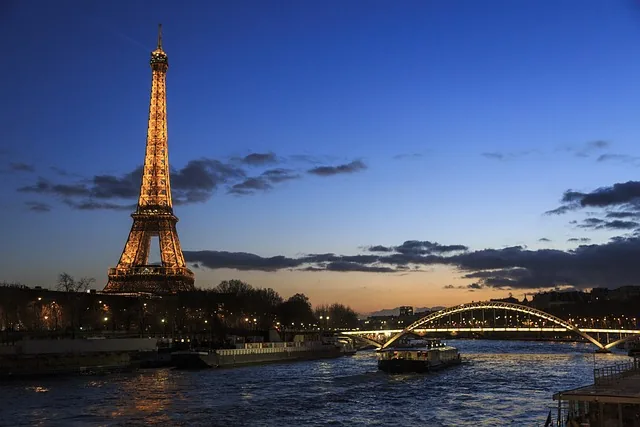 Illuminated Eiffel Tower and lit bridge over the Seine River at dusk, with boats on the water.