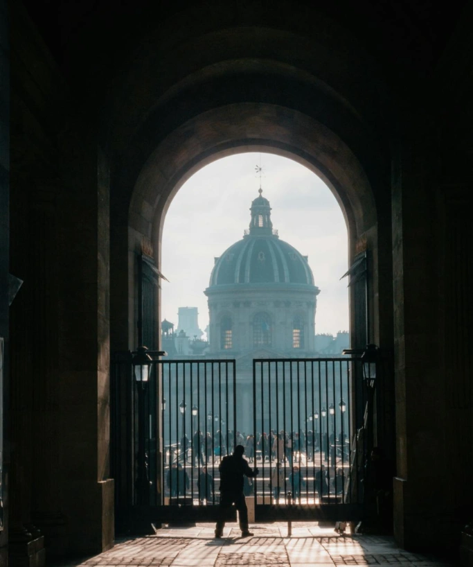 View of the Institut de France dome through a dark stone archway and black metal gate with a silhouetted figure and crowd.