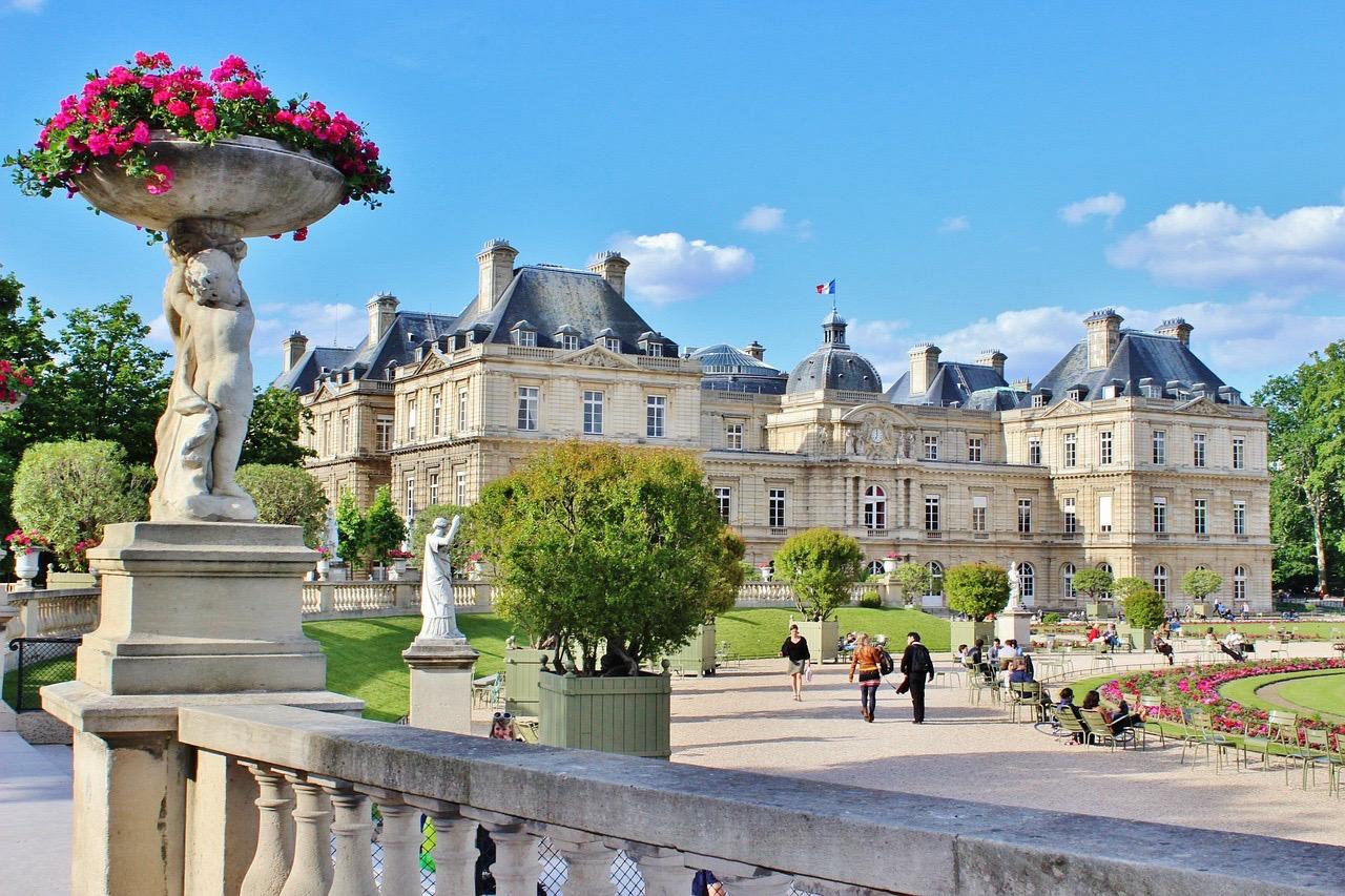 Blick auf den Palais du Luxembourg von den Gärten aus, mit Balustrade, Statuen, blühenden Pflanzen und Spaziergängern.