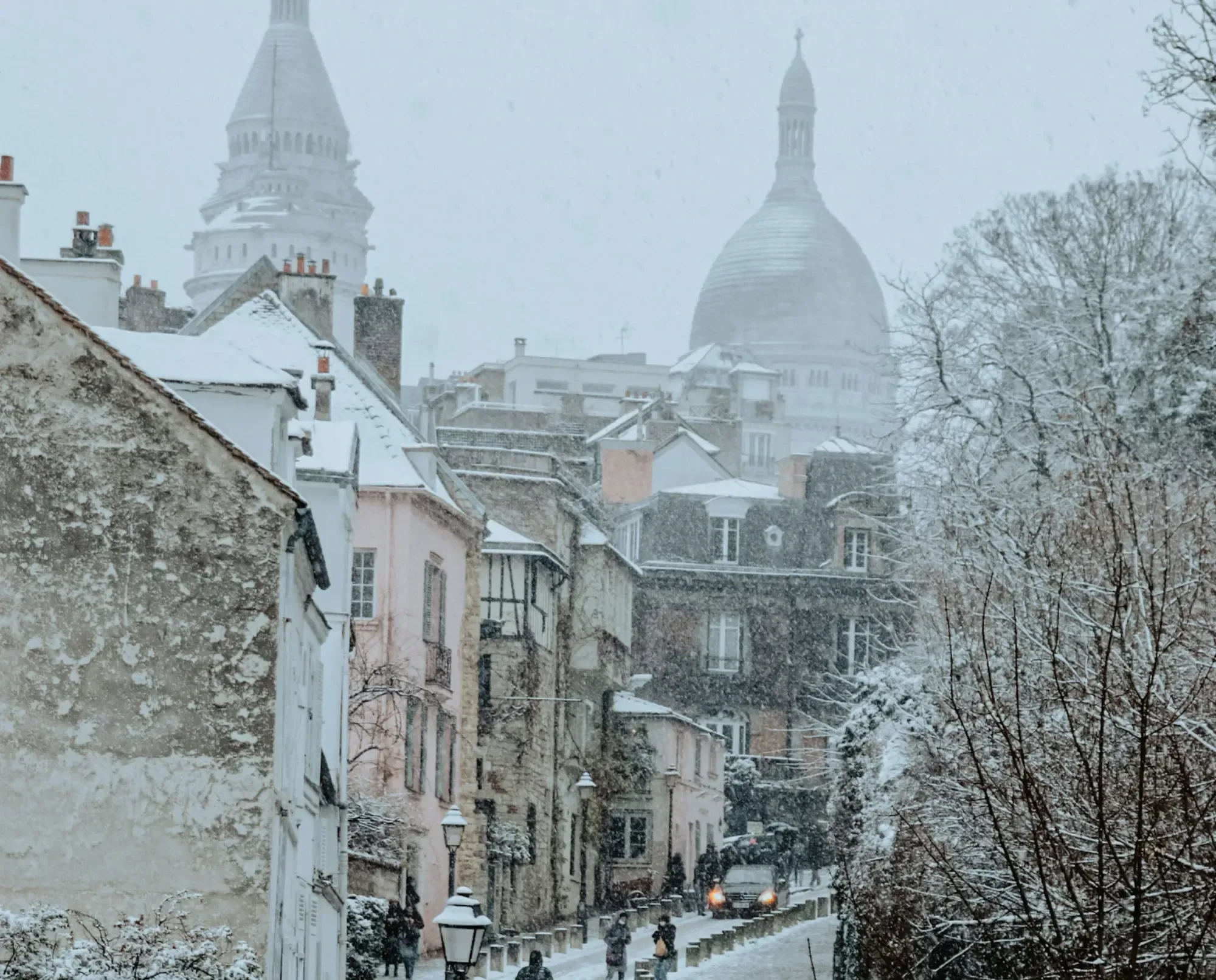 Snowy street view in Montmartre, Paris, with traditional buildings, bare trees, and the Sacré-Cœur Basilica domes in the back