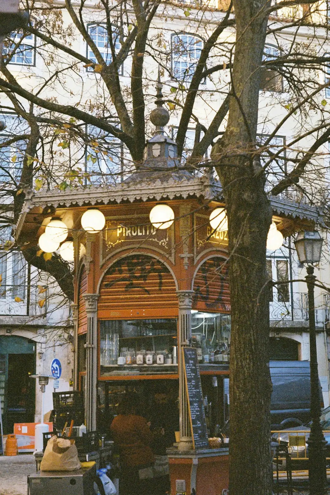 An ornate, old-fashioned street kiosk with round lights, seen through the bare branches of a tree in winter.