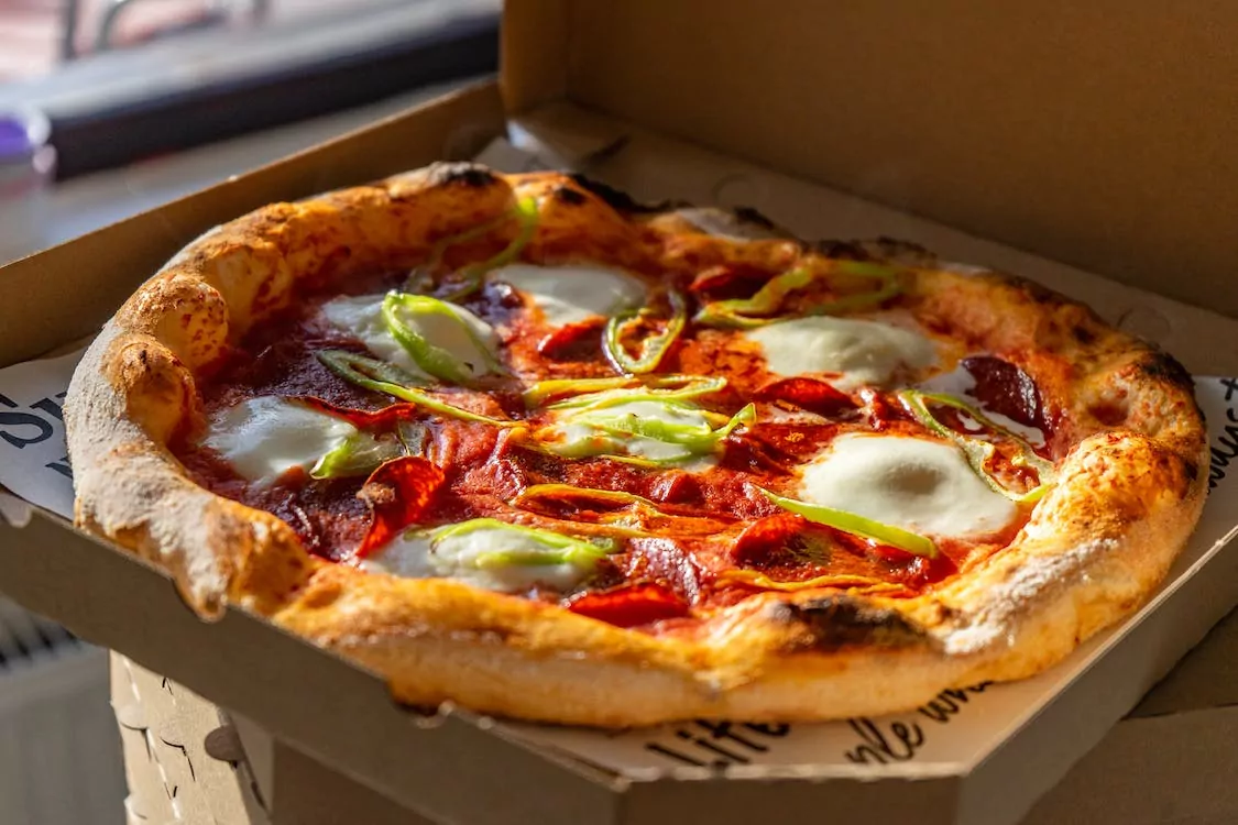 Freshly baked pizza with tomato sauce, pepperoni, mozzarella cheese, and sliced green peppers in an open cardboard delivery b