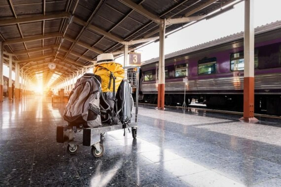 Luggage cart with backpacks and a suitcase on a train station platform next to a purple train, sunlight in the distance.
