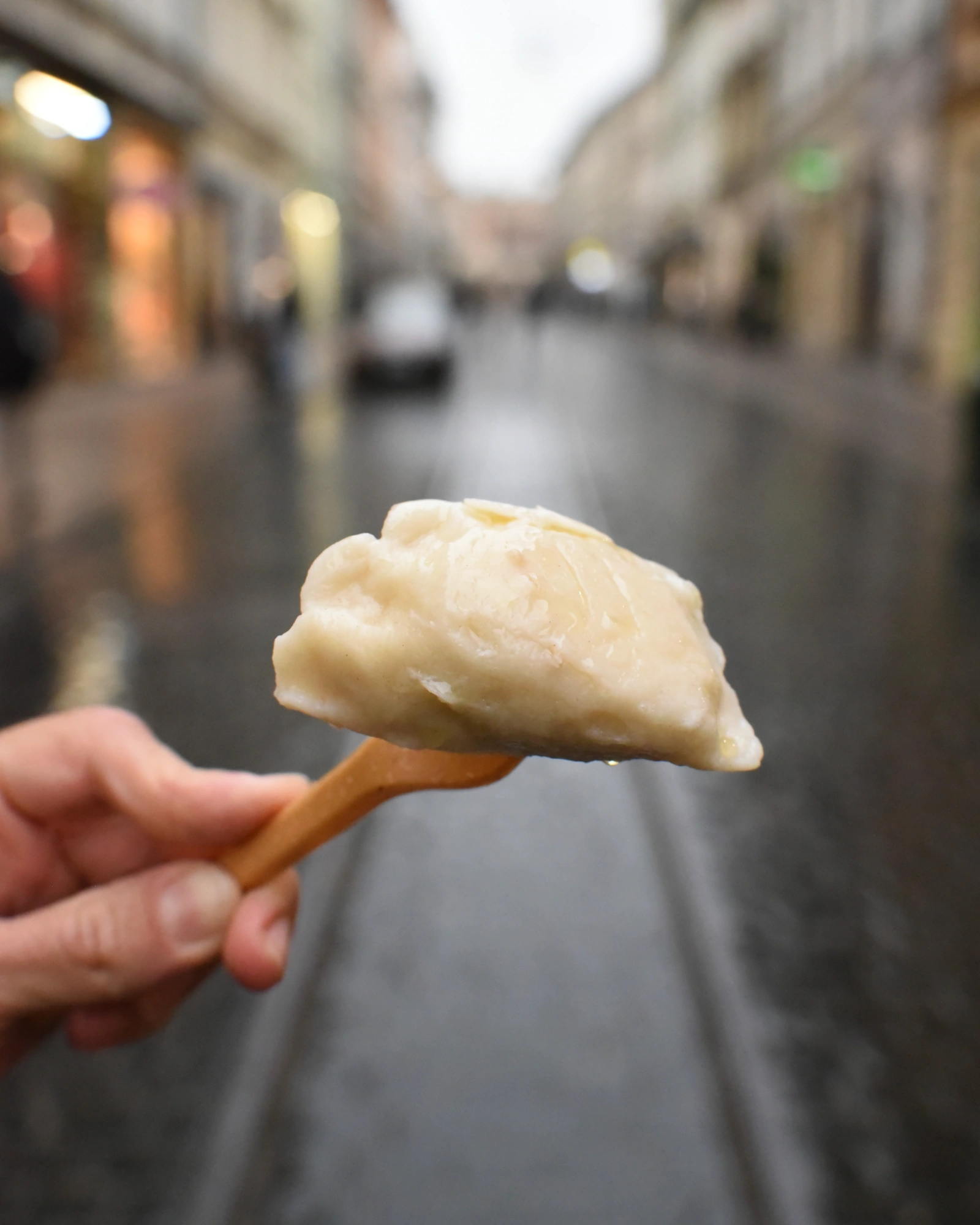 Mano sosteniendo un pierogi polaco en un tenedor de madera con una calle de Cracovia borrosa al fondo.