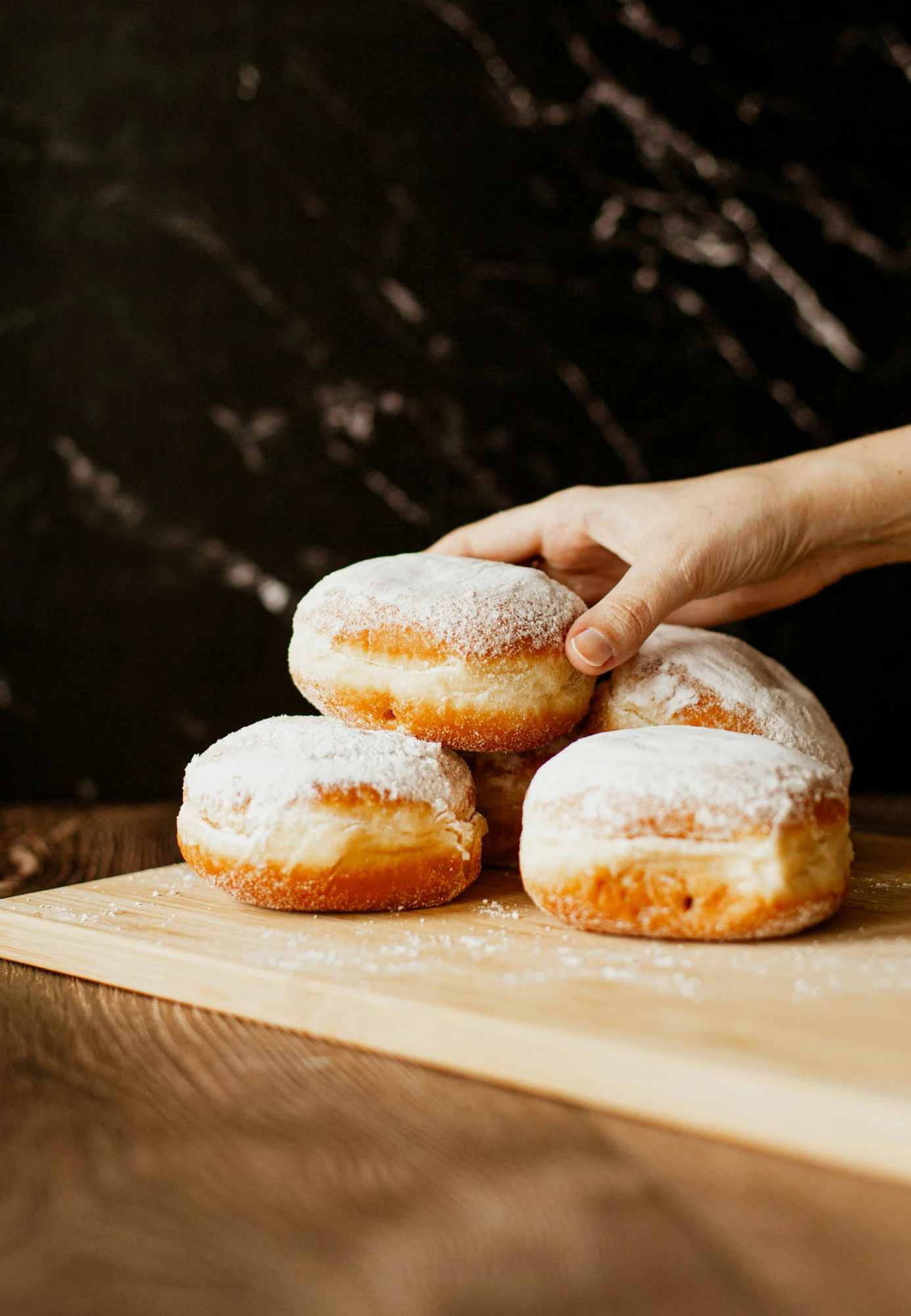 Una mano toma un pączek espolvoreado con azúcar de un montón de dulces polacos tradicionales sobre una tabla de madera.