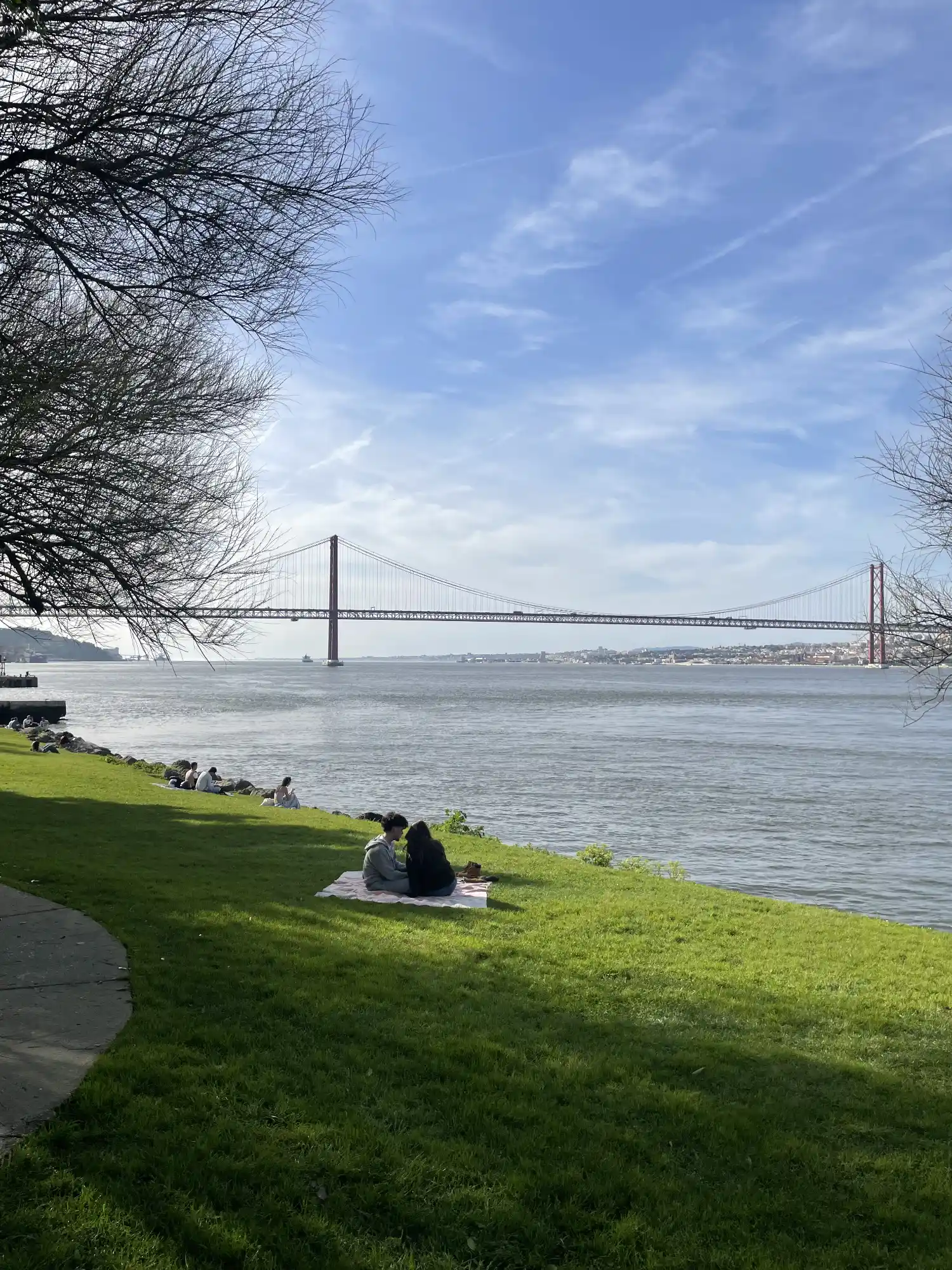 El Puente 25 de Abril cruza el río Tajo. Personas descansan en una orilla verde del río, enmarcada por árboles desnudos bajo