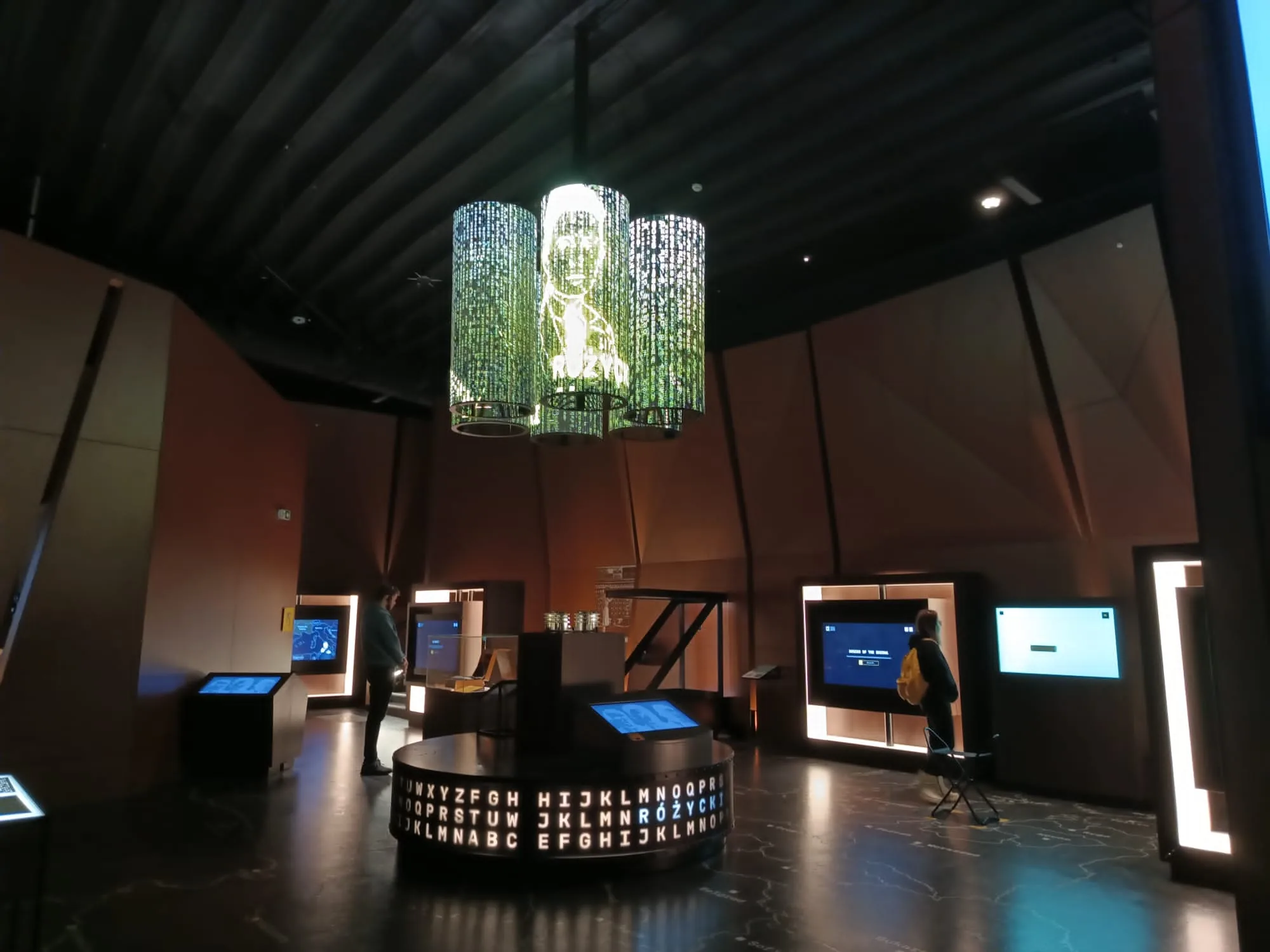 Modern museum interior with interactive exhibits, visitors, and a large round display of letters on the floor.