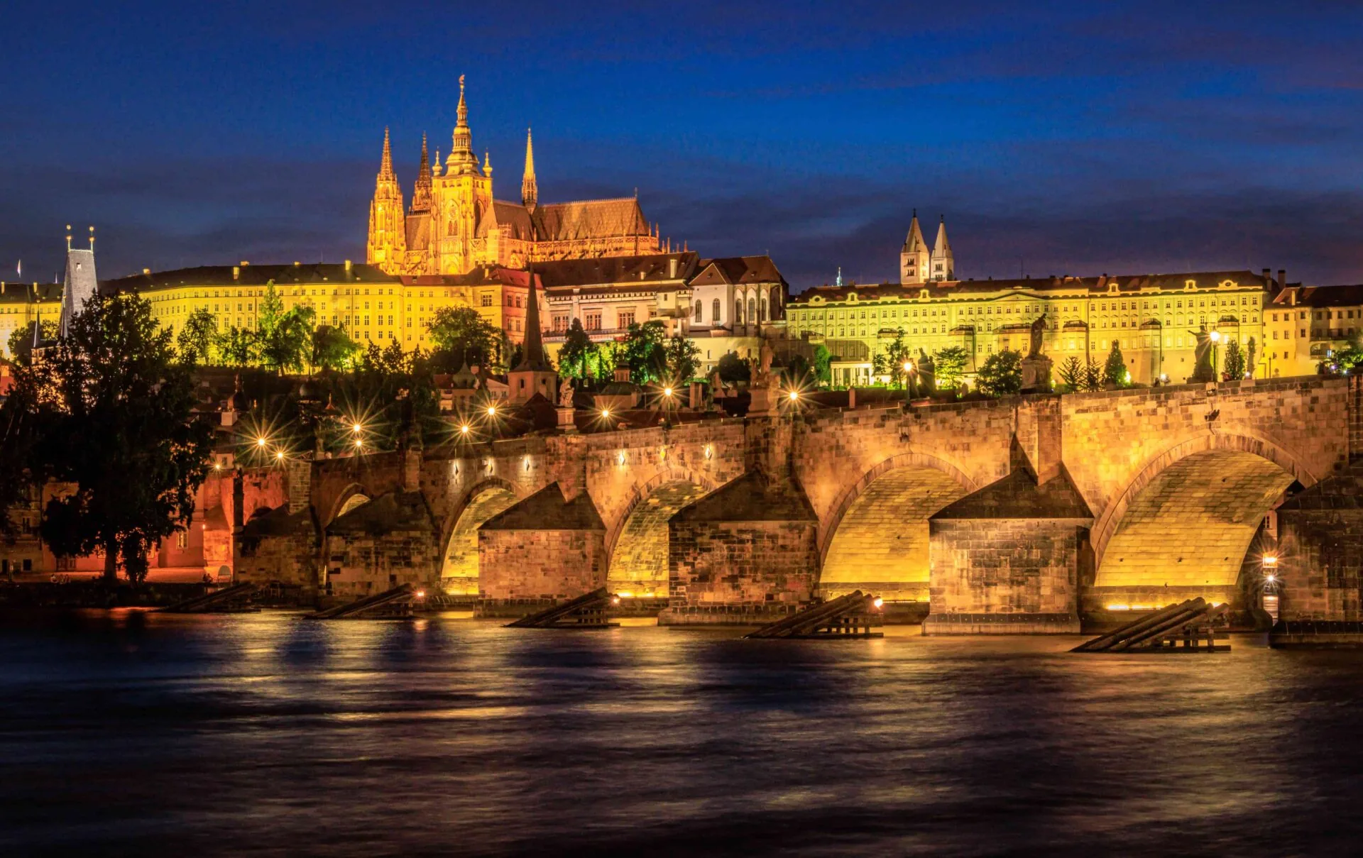 Prague's Charles Bridge and Castle illuminated at night, reflecting in the Vltava River under a dark blue sky.