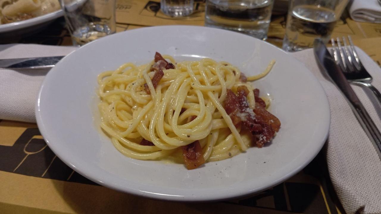Plate of spaghetti carbonara with guanciale, grated cheese, and black pepper on a restaurant table with cutlery and glasses.