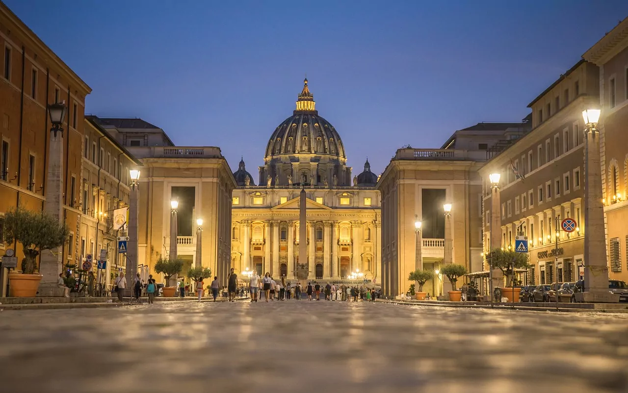 St. Peter's Basilica illuminated at dusk, viewed from a street with pedestrians and buildings on both sides.