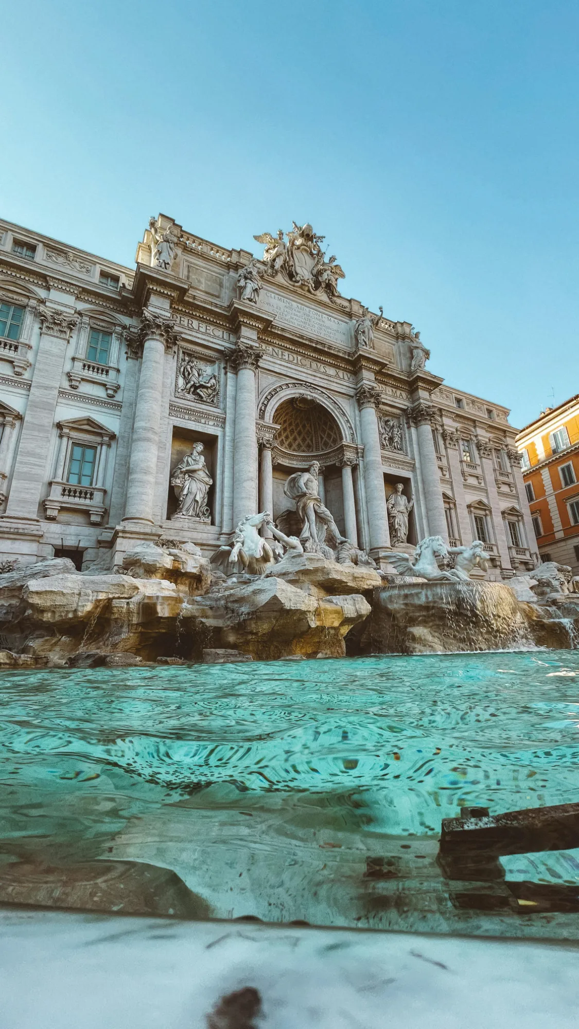 Vista amplia de la Fontana di Trevi en Roma, mostrando su fachada ornamentada, esculturas y agua azul clara con reflejos.