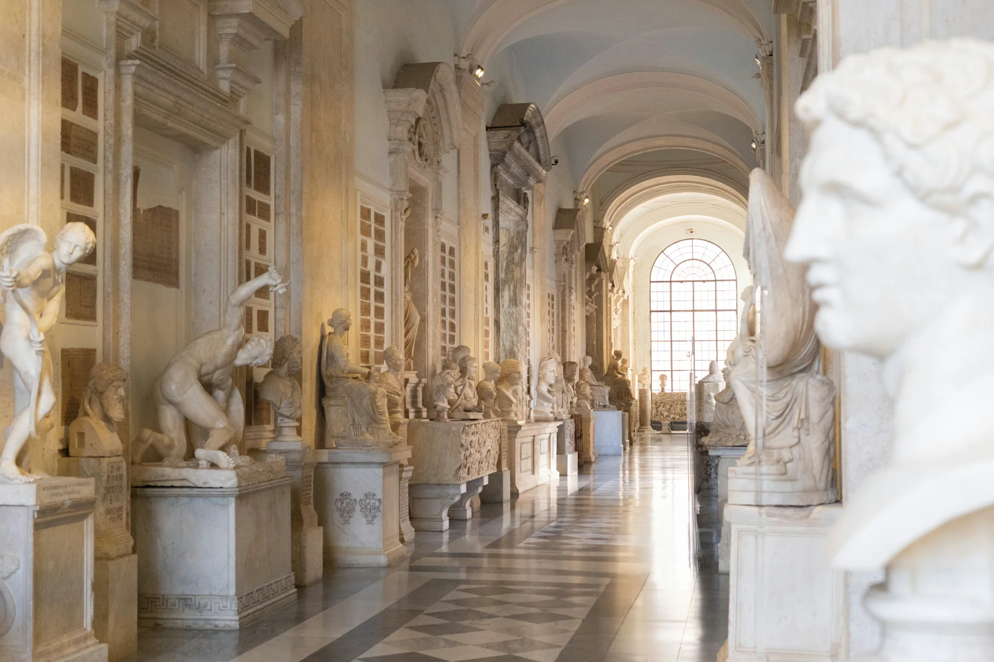 A long, sunlit museum gallery lined with ancient Roman marble statues and busts on pedestals.