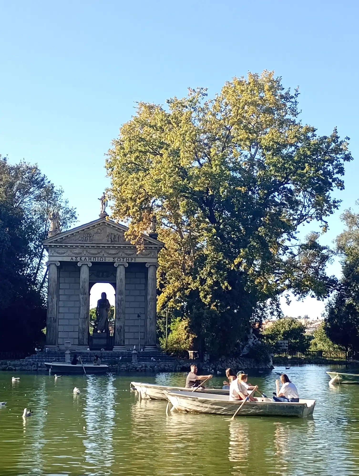 Classical temple with columns and statues by a pond with people rowing boats in Rome's Villa Borghese.