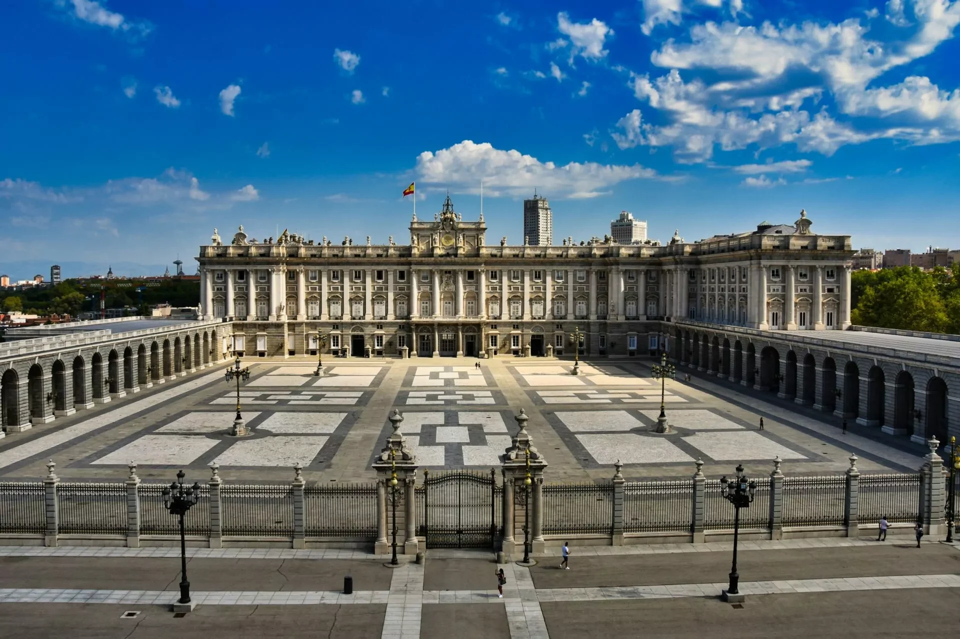 Grand Royal Palace of Madrid with a Spanish flag on its roof, facing a large, patterned stone courtyard. Arcades are visible