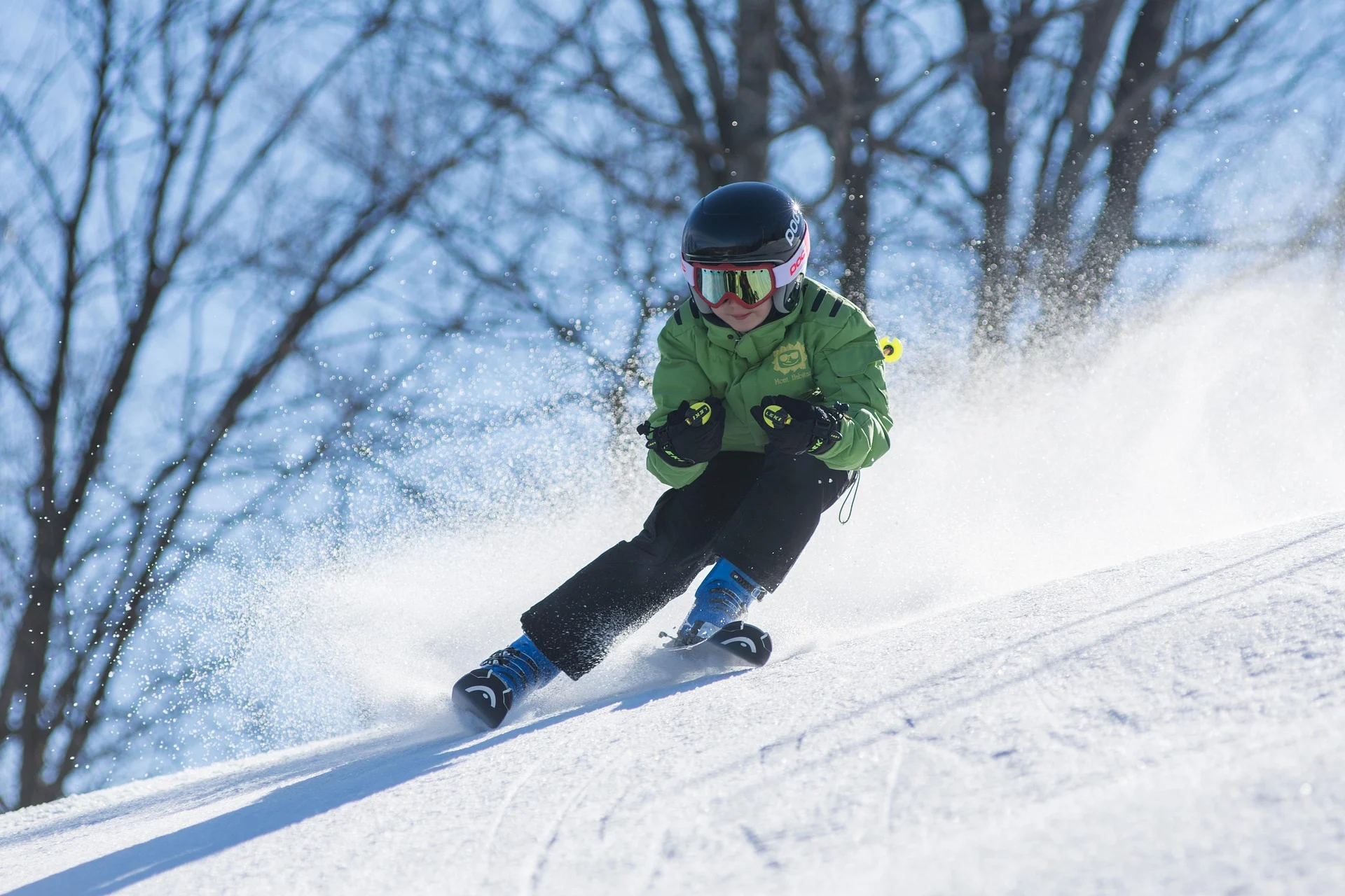 Person in green jacket and black helmet skiing down a sunny snow slope, kicking up snow. Bare trees against blue sky in backg