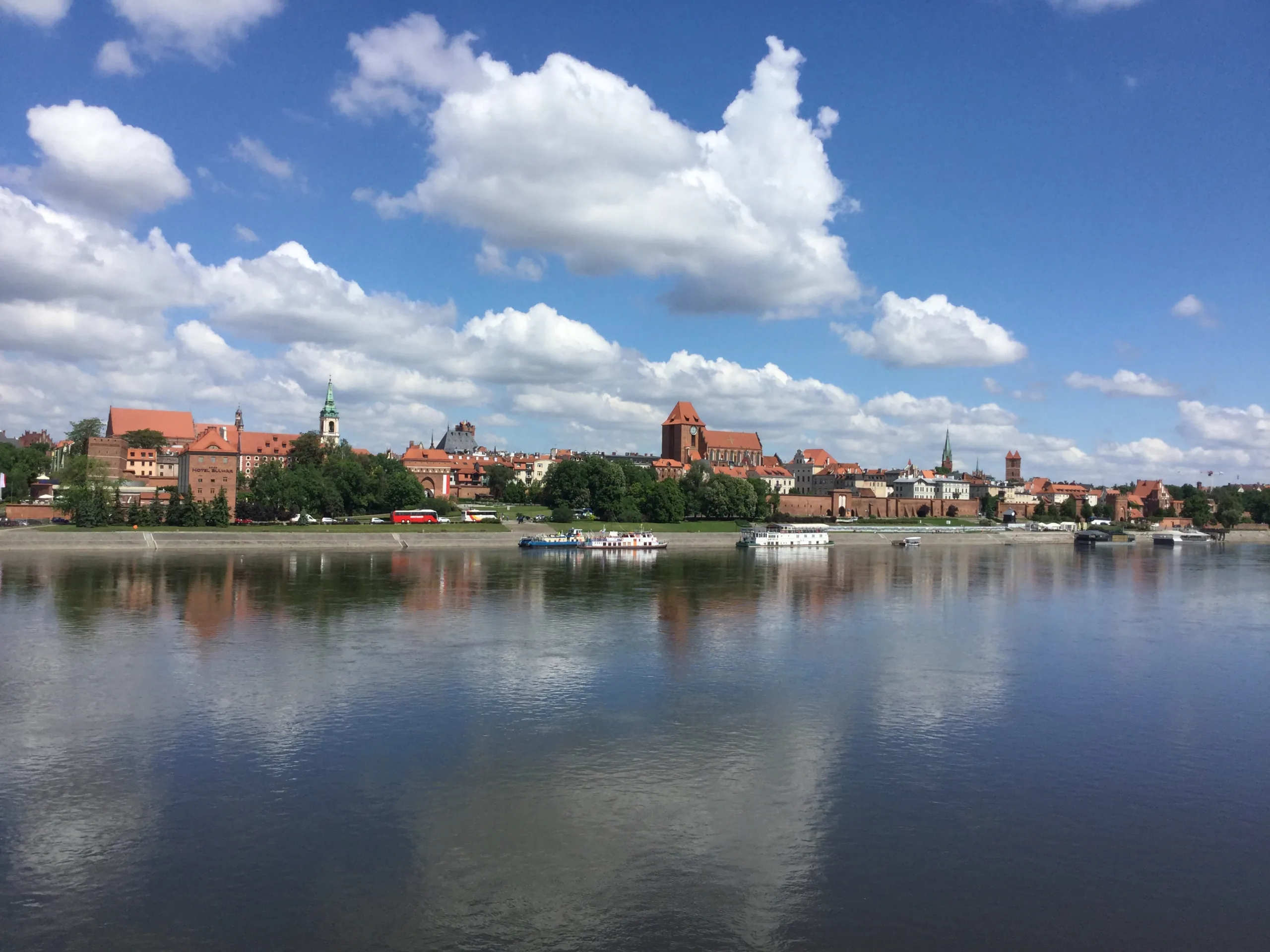 Panoramic view of Torun Old Town skyline across the Vistula River, with historic brick buildings, green trees, and boats on t