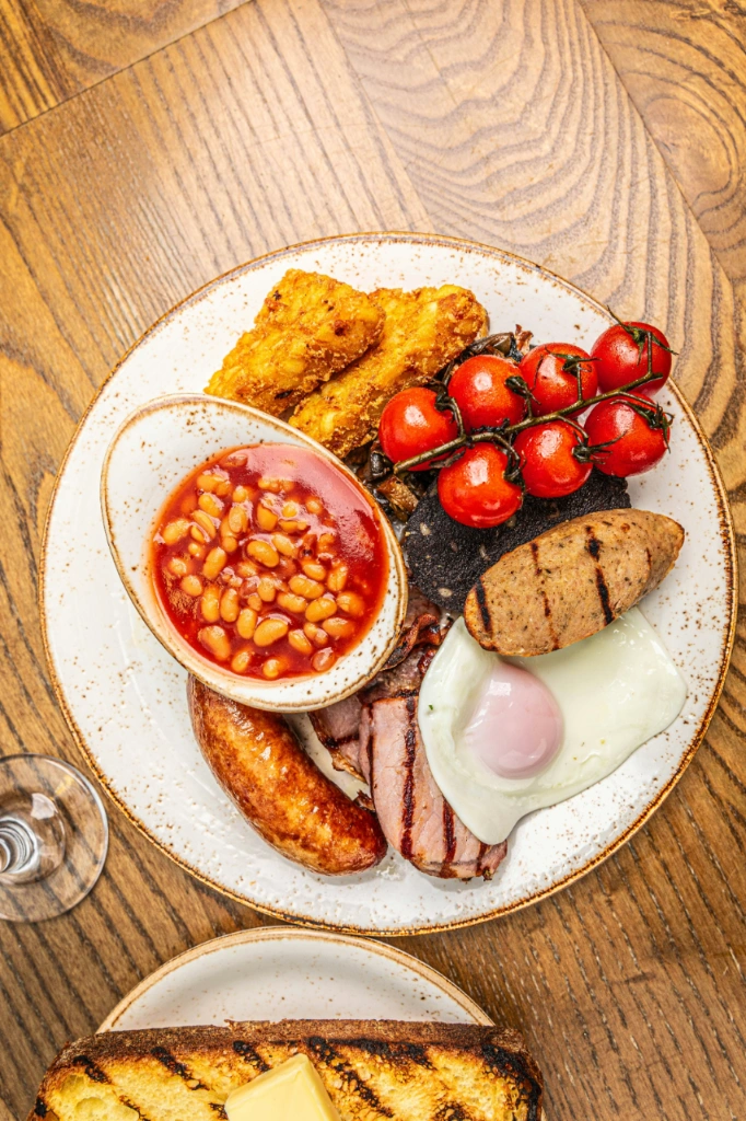Pełne angielskie śniadanie na białym talerzu: fasolka, hash browns, pomidorki koktajlowe, kaszanka, kiełbaska, bekon i jajko