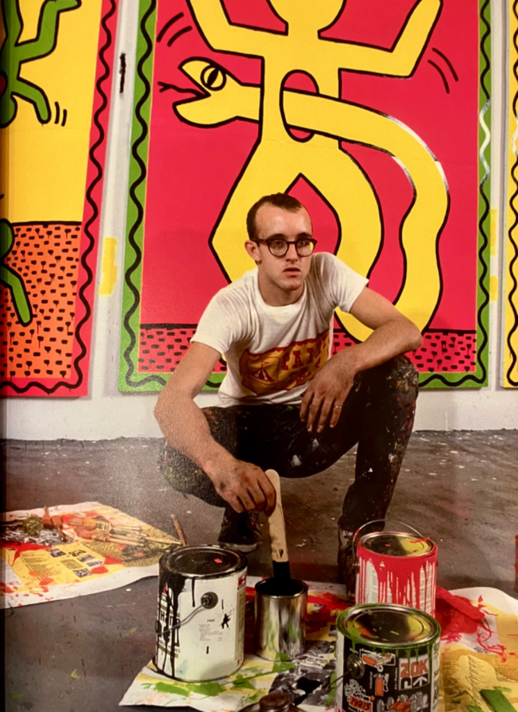 Keith Haring hockt in einem Atelier, umgeben von seinen bunten Pop-Art-Gemälden, Farbdosen und Pinseln.