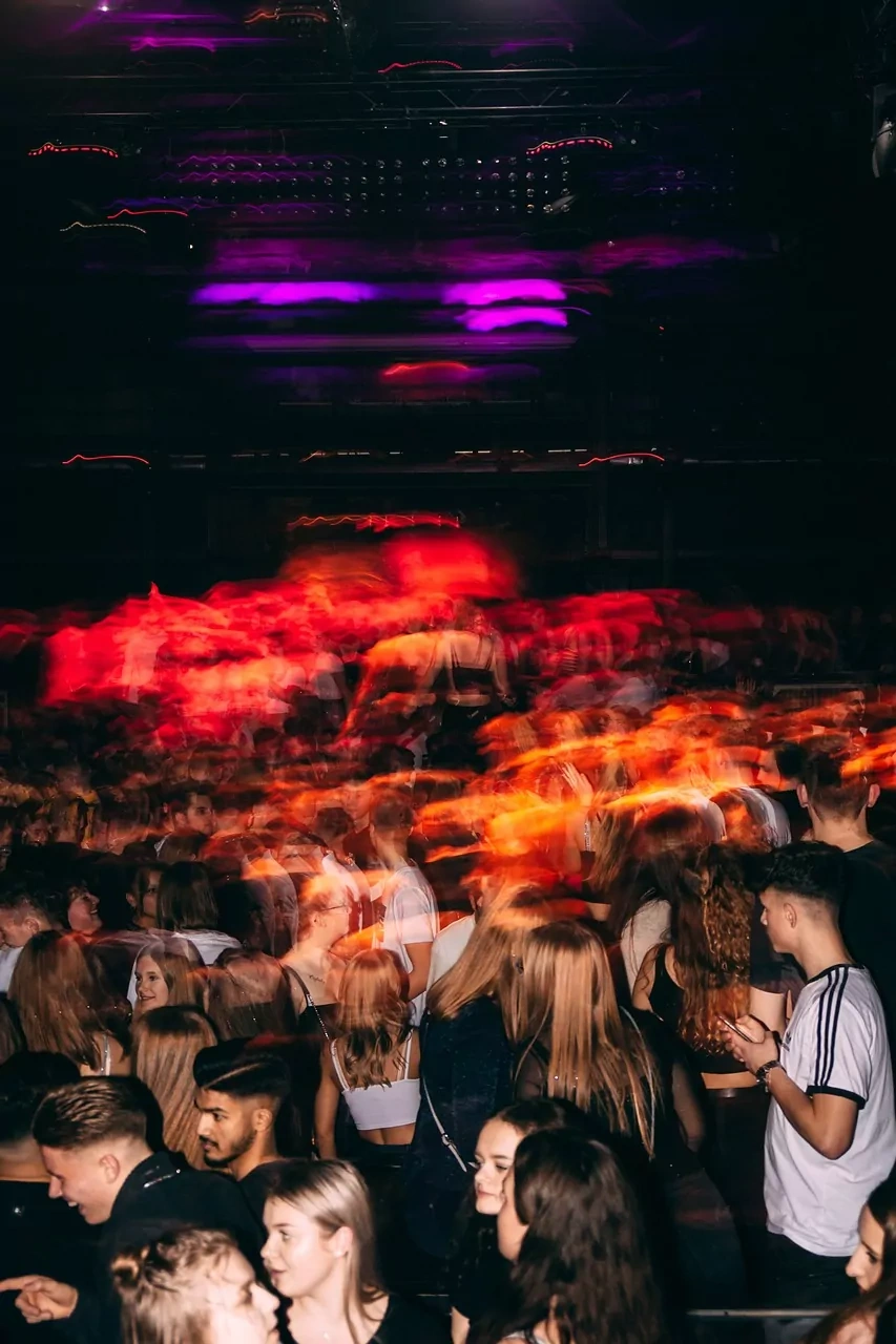 People blurred by motion in a dark nightclub, illuminated by vibrant red and purple lights reflecting on the crowd.