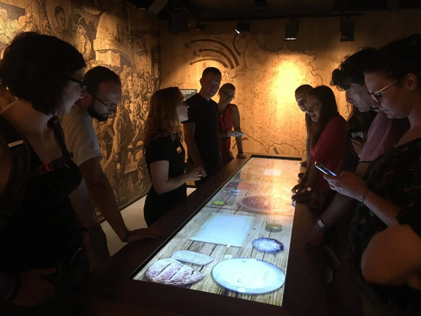 Grupo de personas observando una mesa expositora interactiva iluminada con gráficos de comida en una sala de museo con poca l