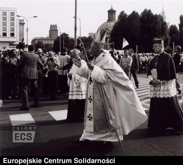Pope John Paul II in full vestments walks across a street during a procession in Warsaw, surrounded by people.
