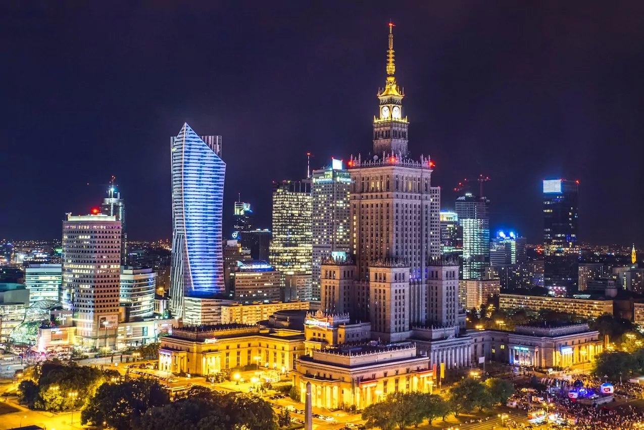 Panorama Warszawy nocą z oświetlonym Pałacem Kultury i Nauki, nowoczesnymi drapaczami chmur i światłami miasta.