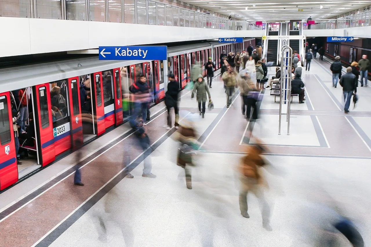 People moving on a modern, brightly lit metro station platform in Warsaw, with a red and white train stopped at the tracks.