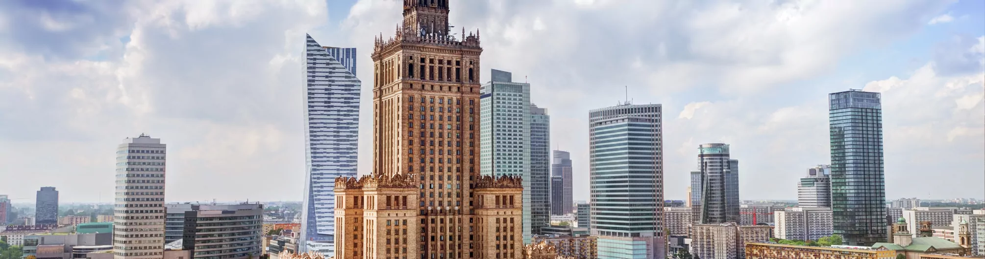 Widok na Pałac Kultury i Nauki, wysoki brązowy budynek, otoczony nowoczesnymi szklanymi wieżowcami pod zachmurzonym niebem.