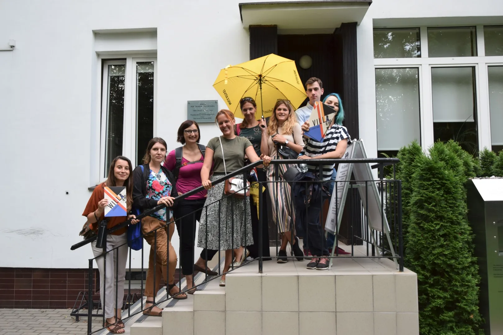 Un grupo de personas de pie en las escaleras exteriores de un edificio blanco. Algunos sostienen libros; un paraguas amarillo