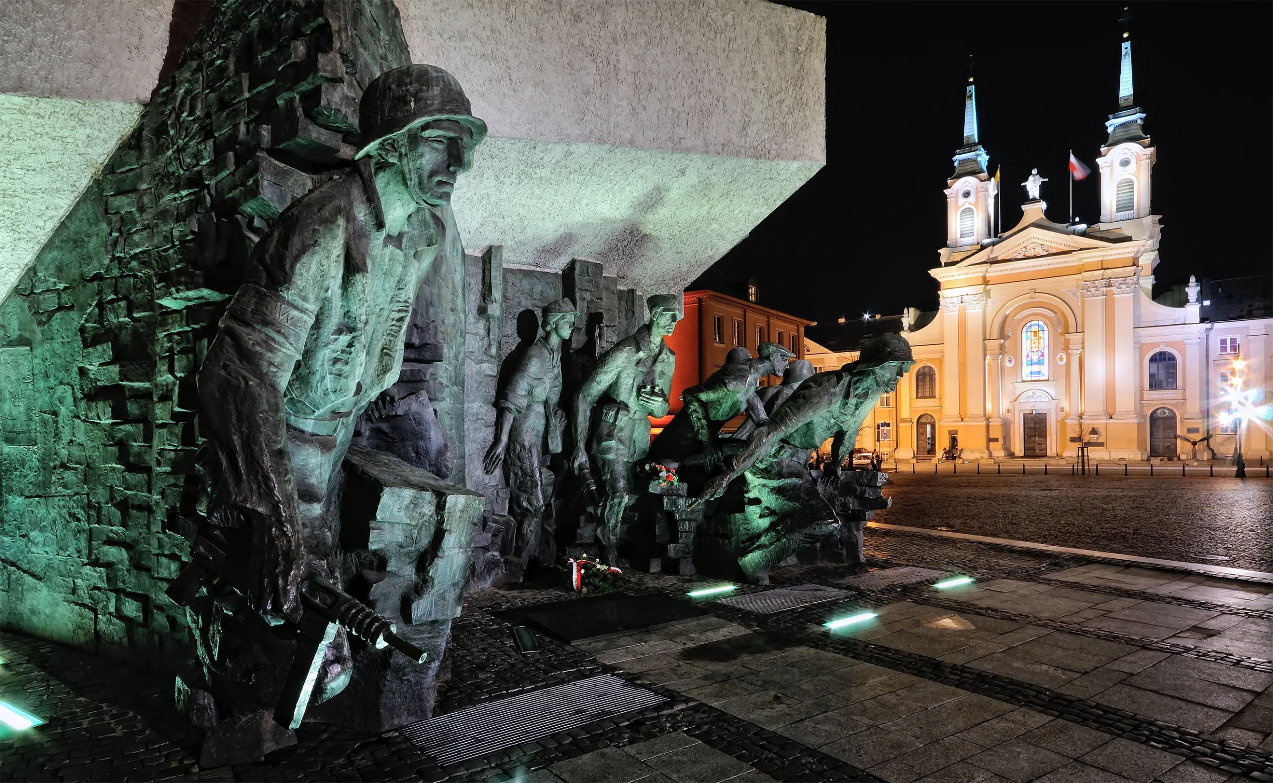 Bronze monument depicting figures of Warsaw Uprising soldiers under green light at night, with a brightly lit church behind.