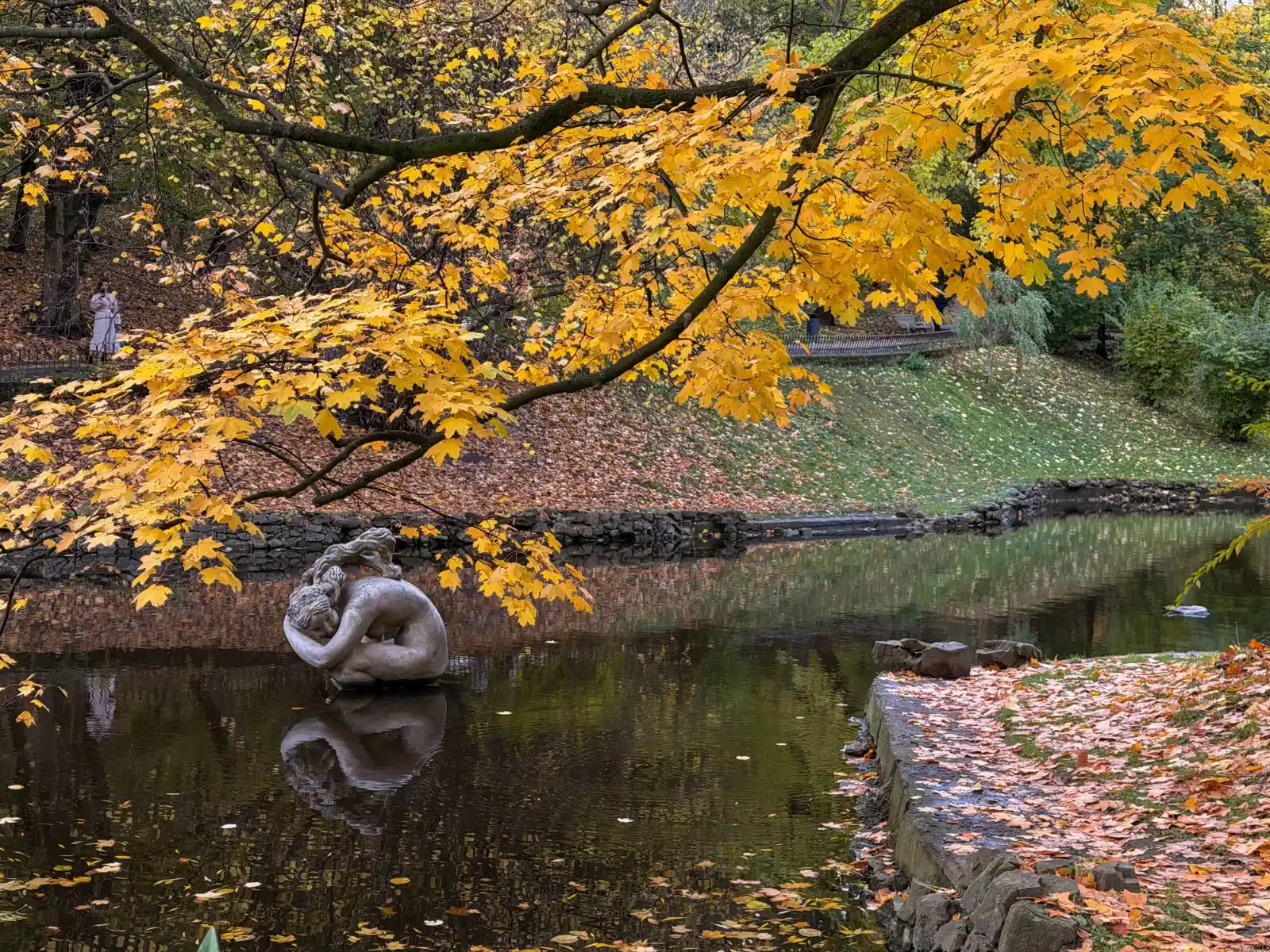 Stone sculpture of a crouching woman in a park pond beneath golden autumn maple branches.