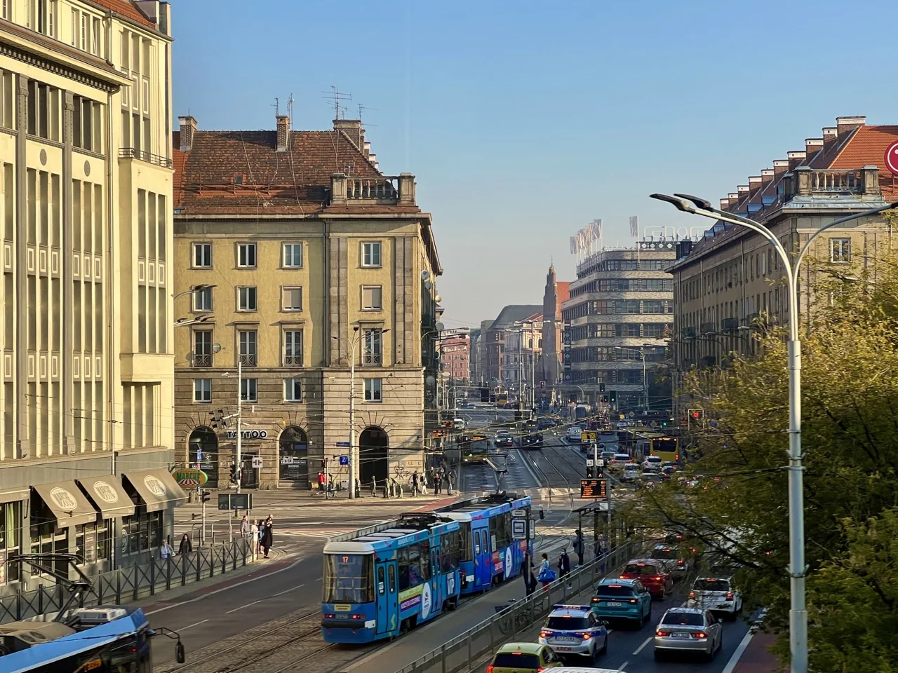 Szeroka ulica miejska z tramwajami, samochodami i pieszymi, otoczona wielopiętrowymi budynkami pod czystym niebem.