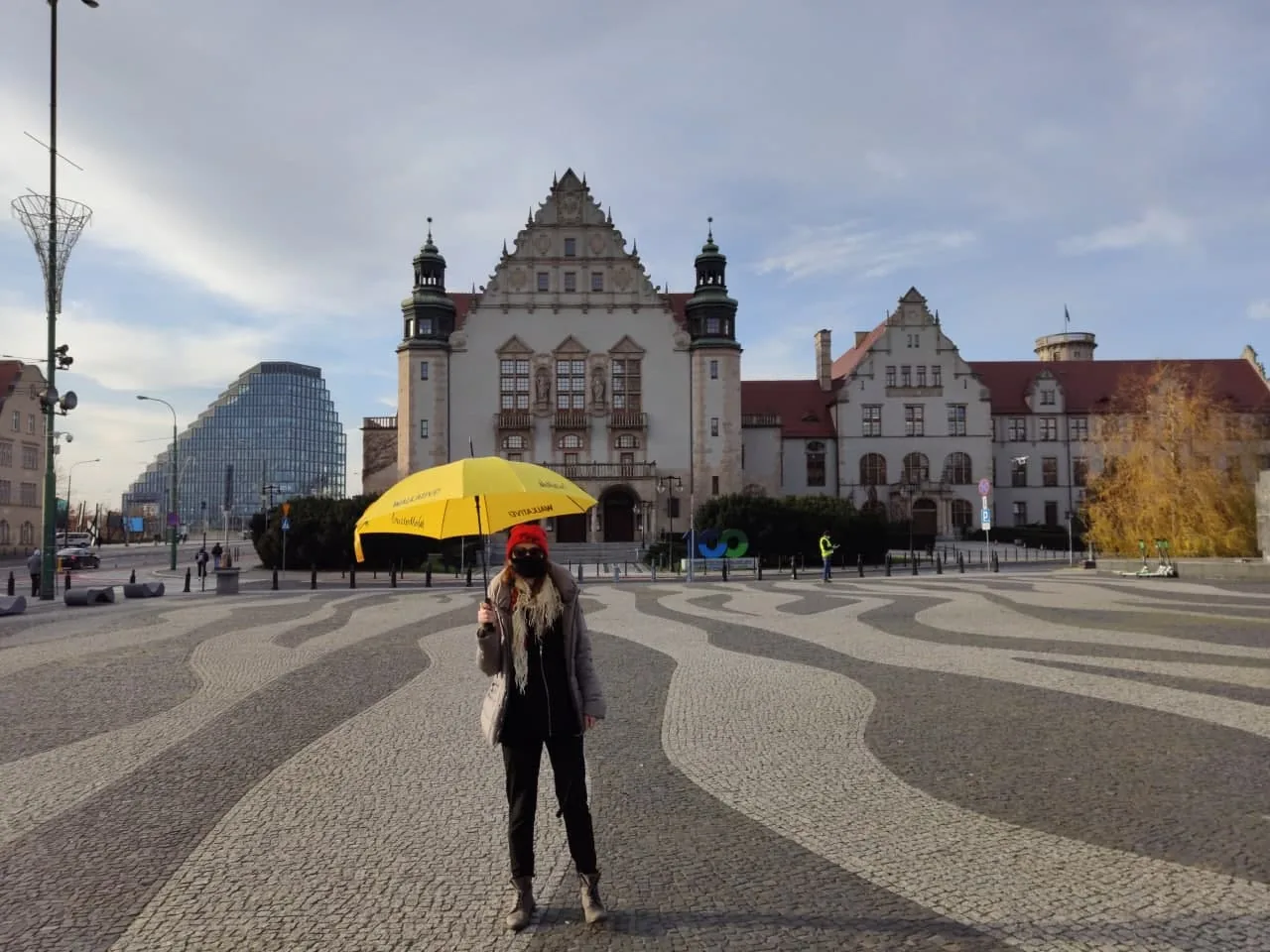 Person mit gelbem Walkative-Regenschirm und Maske auf gepflastertem Platz, mit historischen und modernen Gebäuden im Hintergr