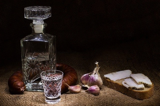 Clear glass decanter and shot glass with vodka, a sausage ring, garlic, and slices of salo on bread, all on burlap.