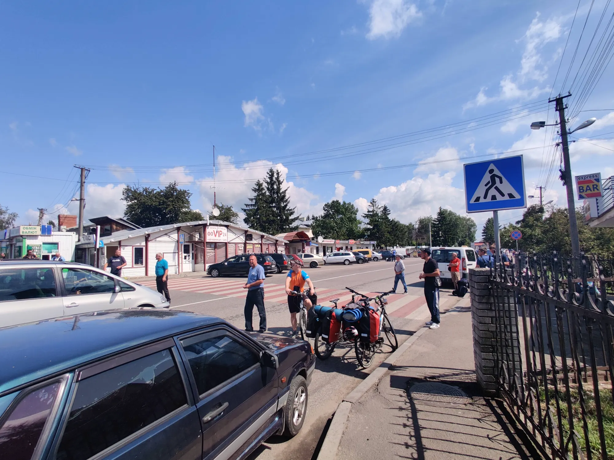 Pedestrians and cyclists waiting at the Shehyni border crossing between Ukraine and Poland on a sunny day.