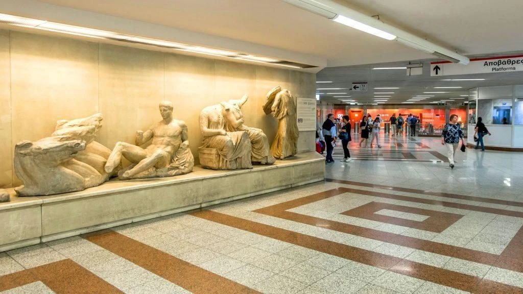 Classical Greek sculpture casts exhibited along the wall of a brightly lit Athens metro concourse.