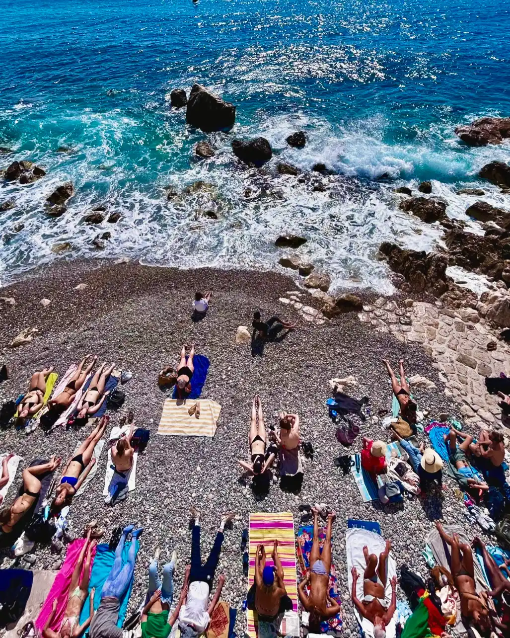 People sunbathe on colorful towels on a pebble beach next to the bright blue sea, seen from a high angle.