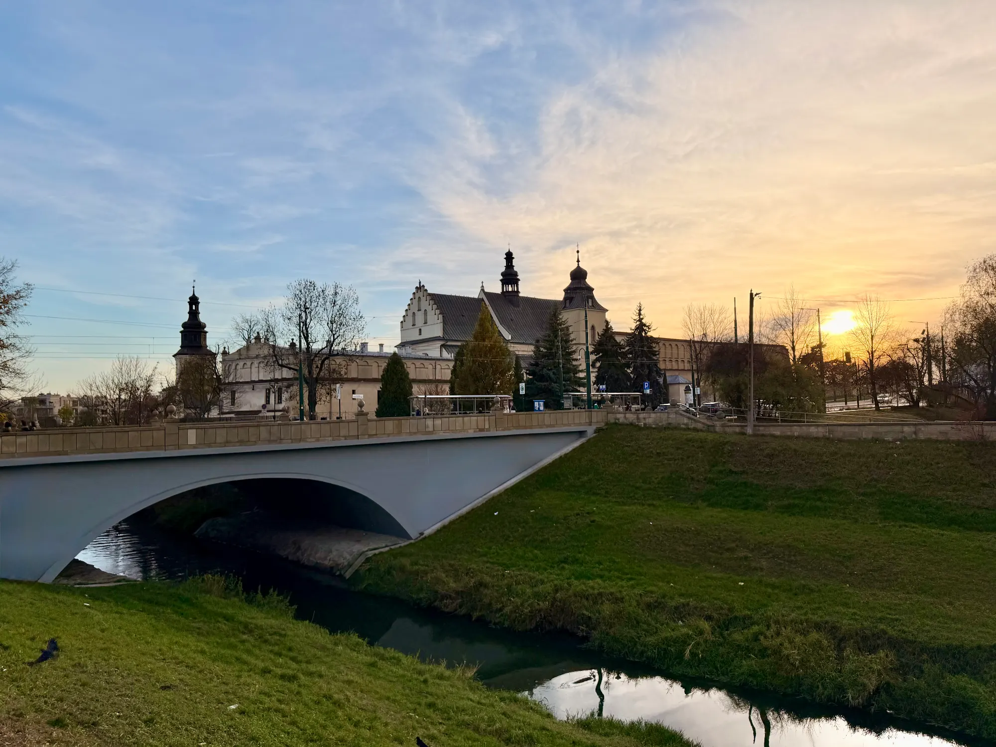 A bridge crosses a river in Krakow, with the Church of St. Catherine and Margaret in the background at sunset.