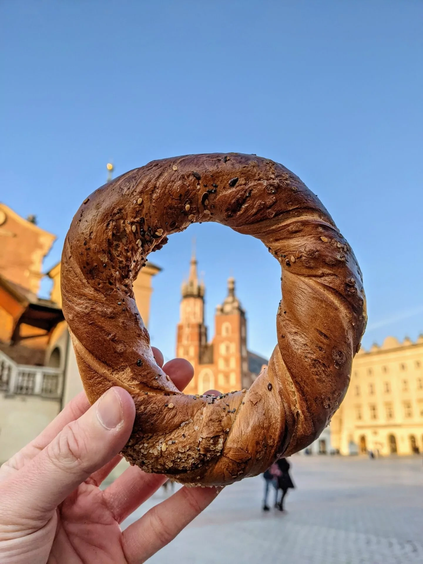 A hand holds a sesame and poppy seed obwarzanek in the foreground, with Krakow's St. Mary's Basilica blurred in the background.