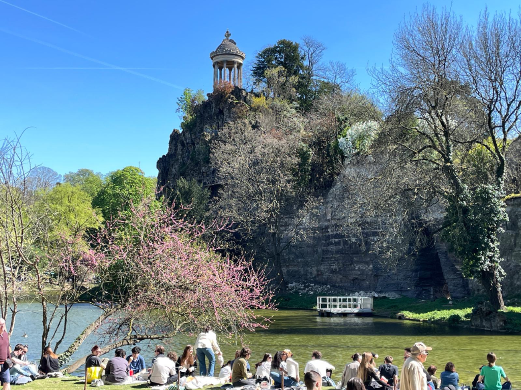 The Temple de la Sibylle stands atop a rocky cliff in Paris's Parc des Buttes-Chaumont, a popular spot for relaxing. People relaxing on grass by a lake in Parc des Buttes-Chaumont, with the Temple de la Sibylle atop a rocky cliff in Paris.