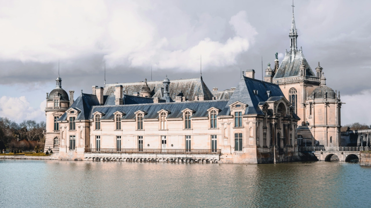 The Chateau de Chantilly in France surrounded by a wide moat under a cloudy sky.