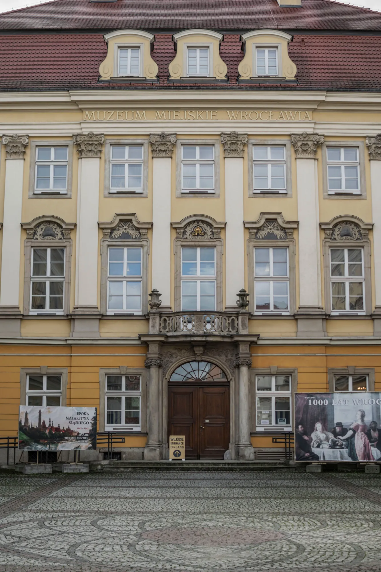 The yellow and white facade of the City Museum of Wroclaw, seen from the cobblestone courtyard in front.