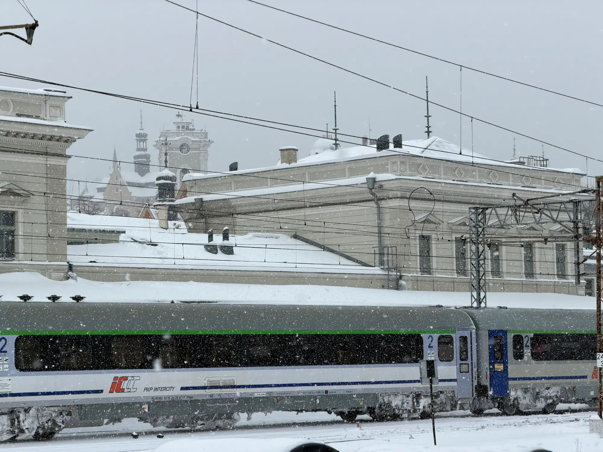 A PKP Intercity train at a snow-covered station in Przemyśl, Poland, with historic architecture in the background.