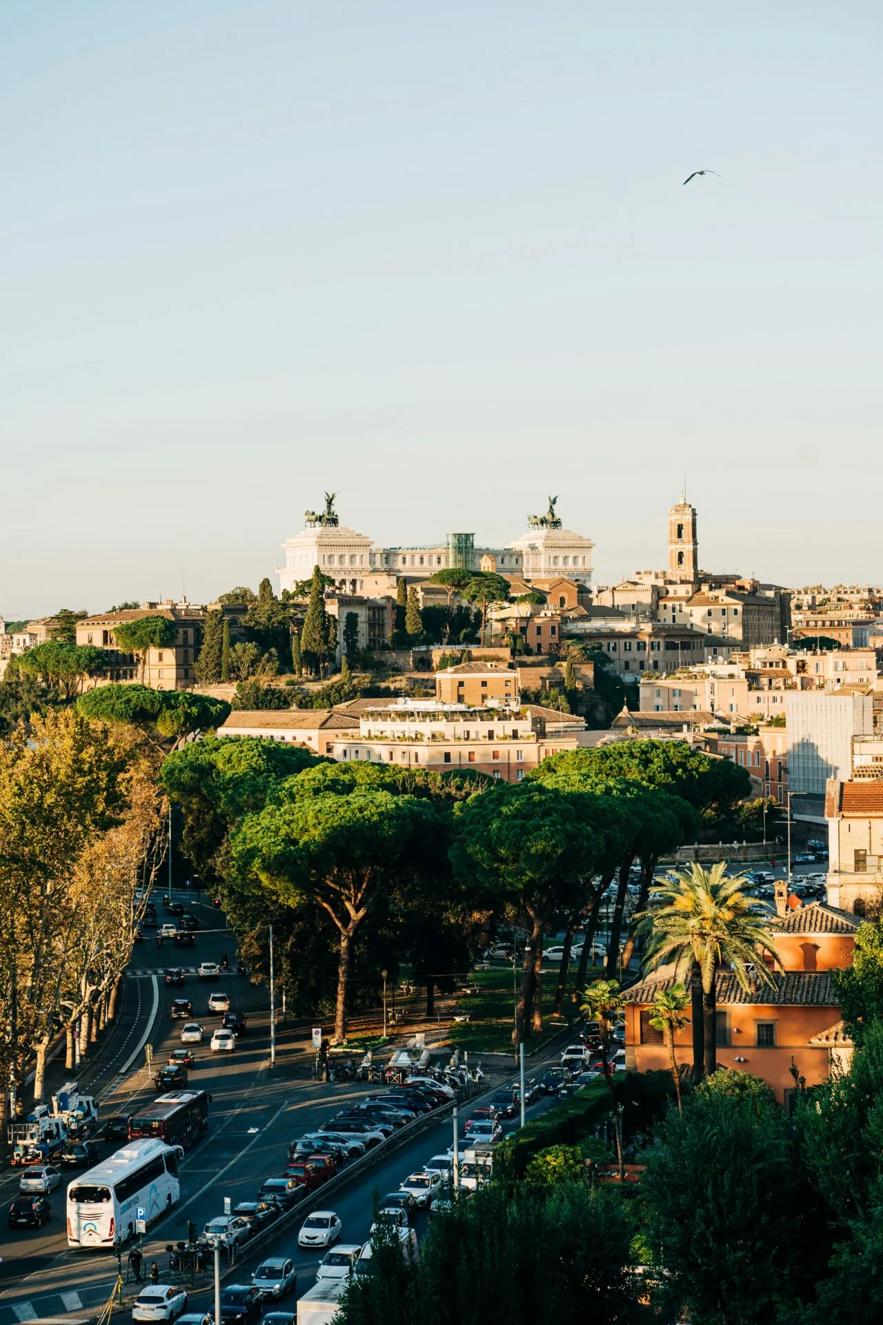 Panoramic view of Rome with the Victor Emmanuel II Monument atop a hill, a busy street with cars and buses, and green trees.