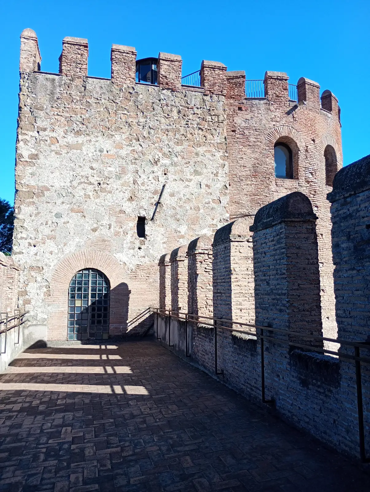 A stone and brick fortified wall with battlements and a paved walkway, under a clear blue sky.