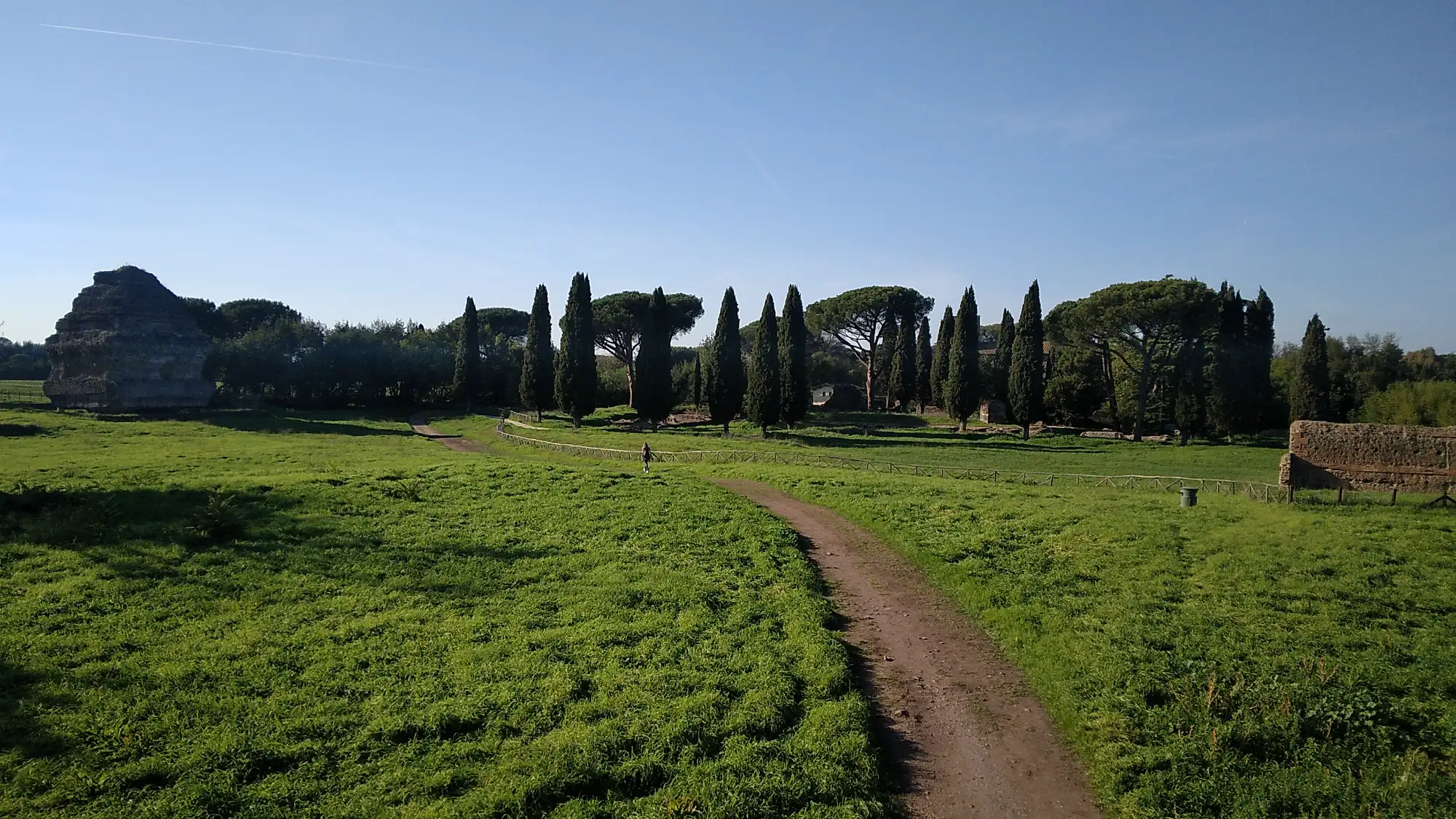 Dirt path winding through a green field with ancient stone ruins and tall cypress trees under a clear blue sky in Rome.