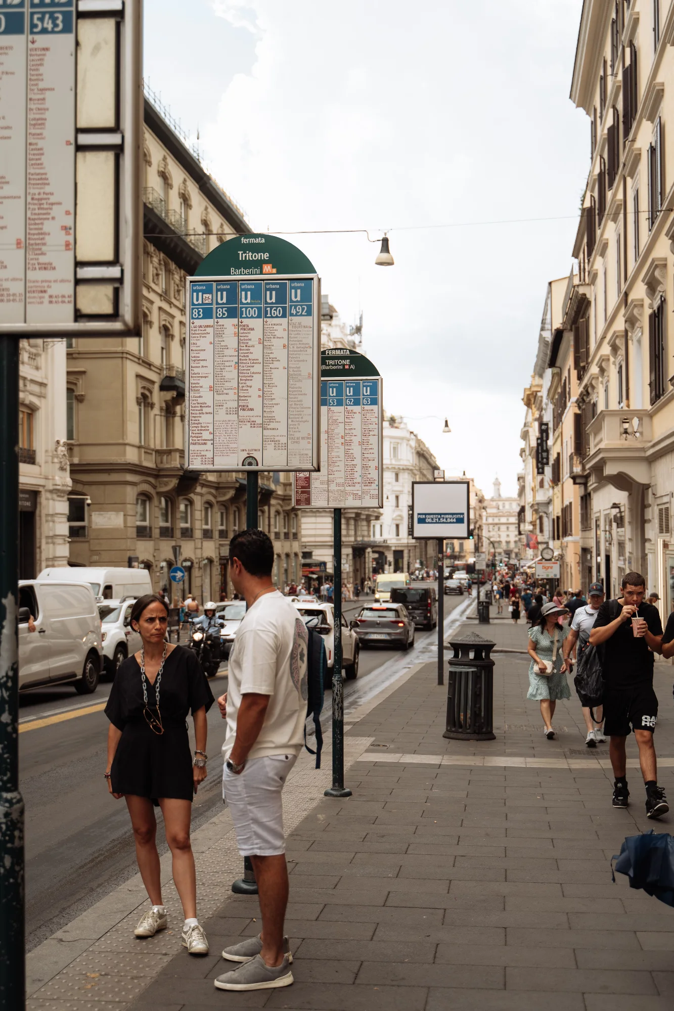 A couple stands at a bus stop on a busy street in Rome, with timetables and city buildings in the background.