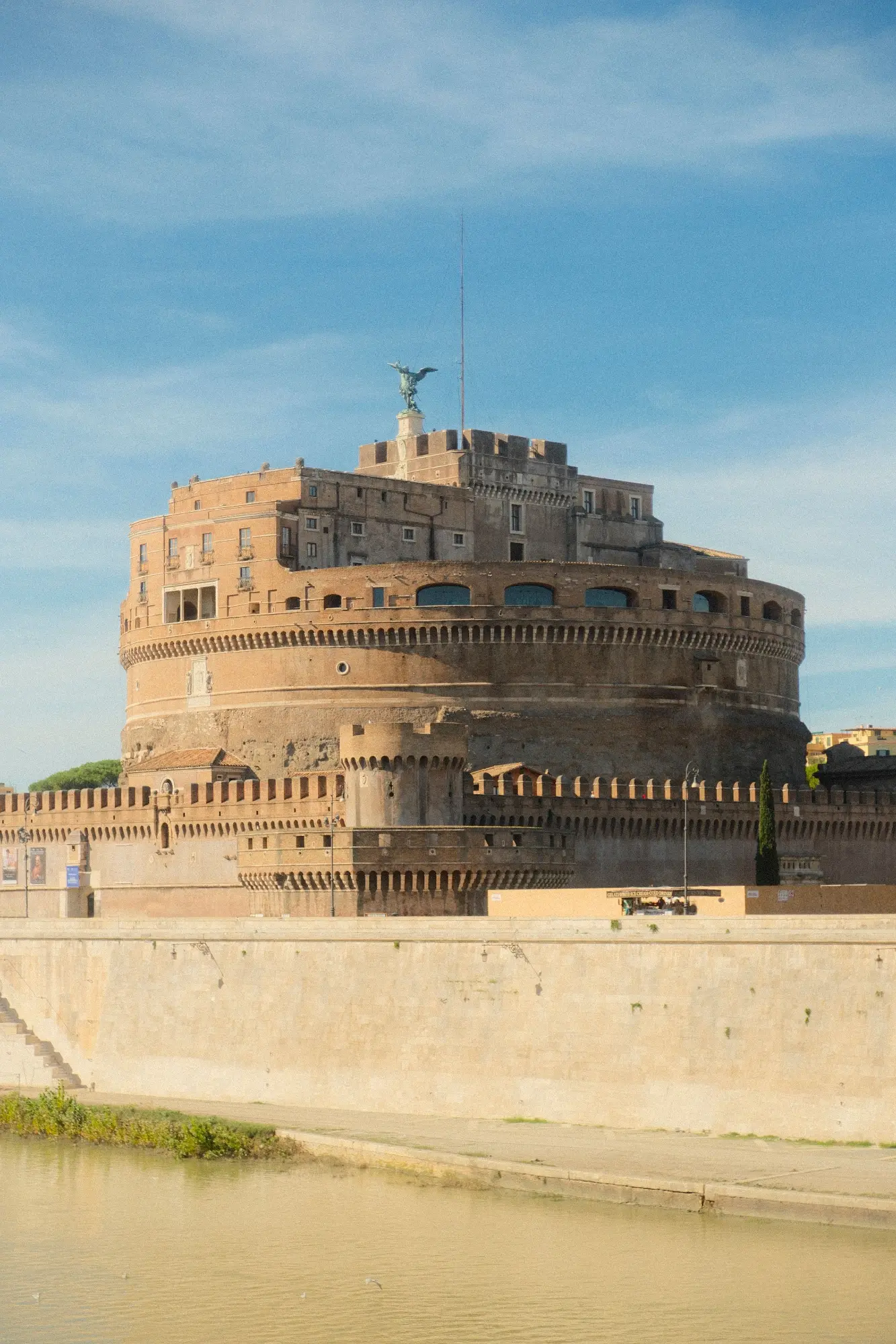 The Castel Sant'Angelo in Rome, a cylindrical stone fortress, seen from across the Tiber River on a clear day.
