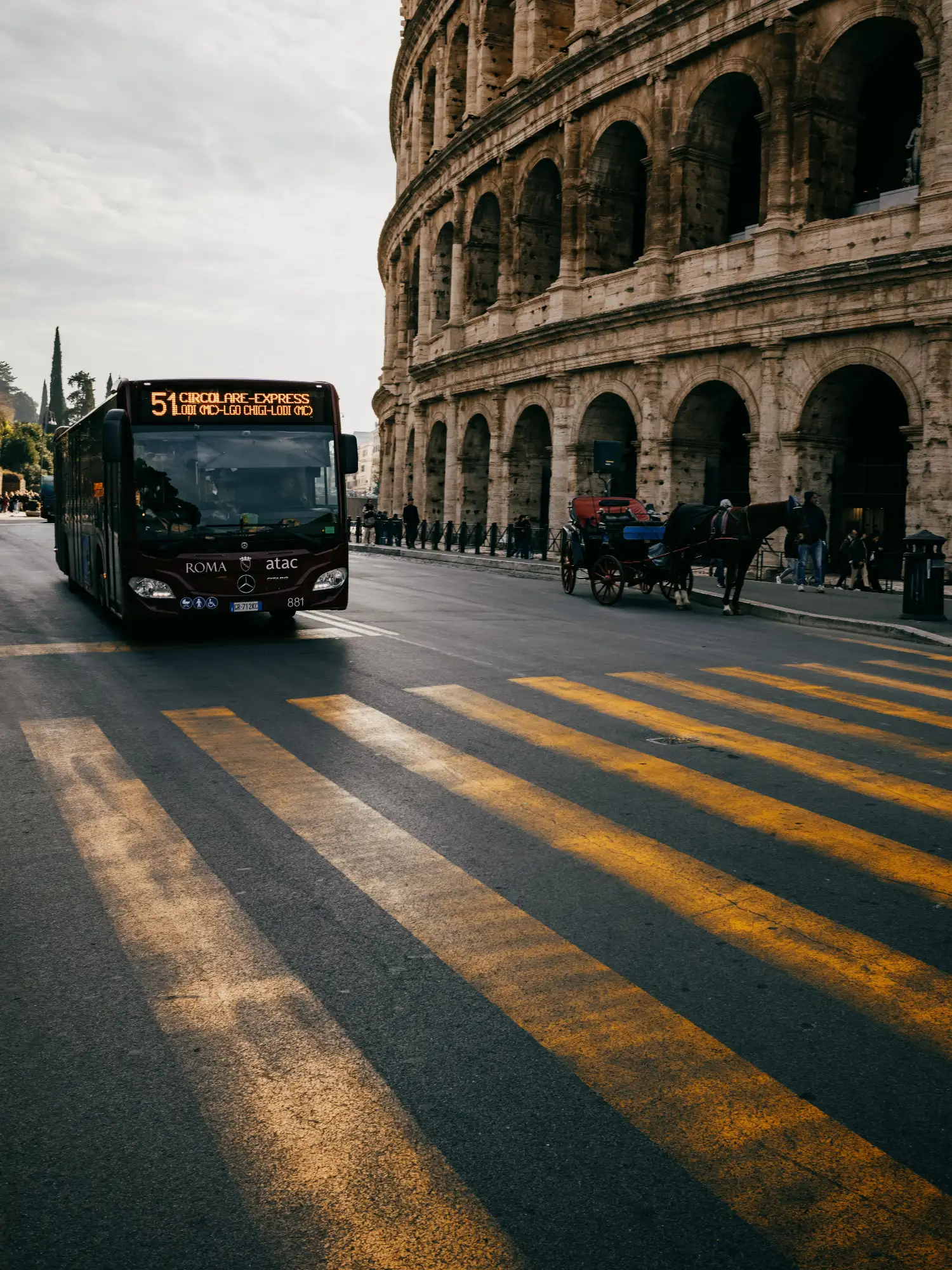 A city bus and a horse-drawn carriage on the street next to the ancient Colosseum in Rome.